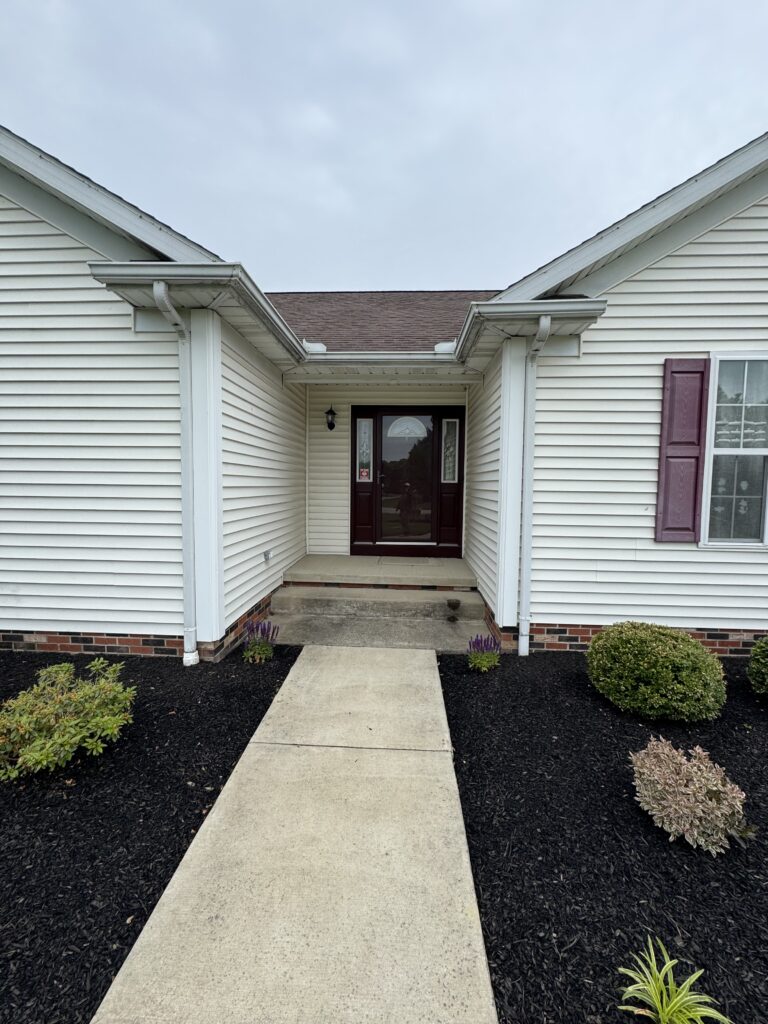 Front entryway of a suburban house: a concrete walkway leads to dark red front doors under a small covered porch, cream siding and dark mulch beds.