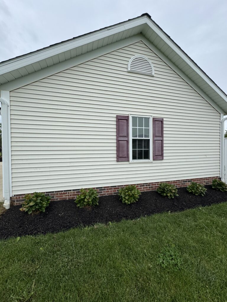 Side view of a beige house with vinyl siding, a window with burgundy shutters, and a brick foundation garden bed with mulch.
