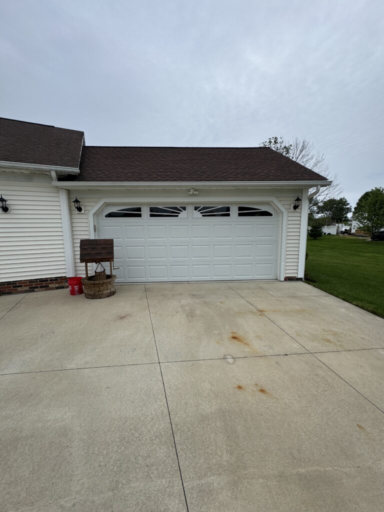 White suburban garage with a brown shingled roof, closed two-car door, and paved driveway; small red bucket and wooden stand on the left.