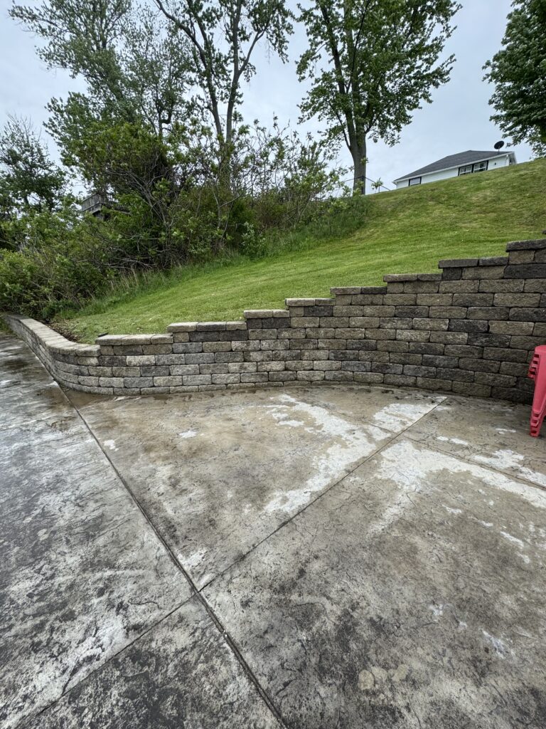 Concrete patio in the foreground with a curved stone retaining wall, a grassy slope, and trees beyond; a white house sits on the hill in the background.