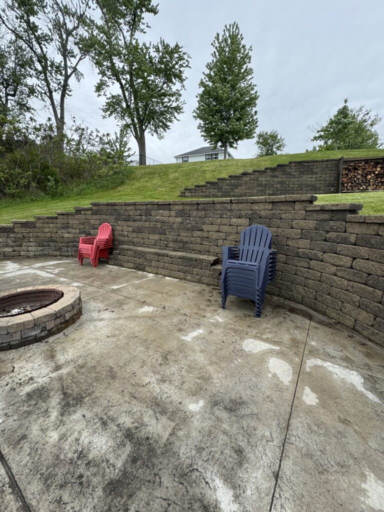 Red and blue plastic chairs sit near a circular fire pit on a concrete patio, with a stacked stone wall and grassy hill behind them.