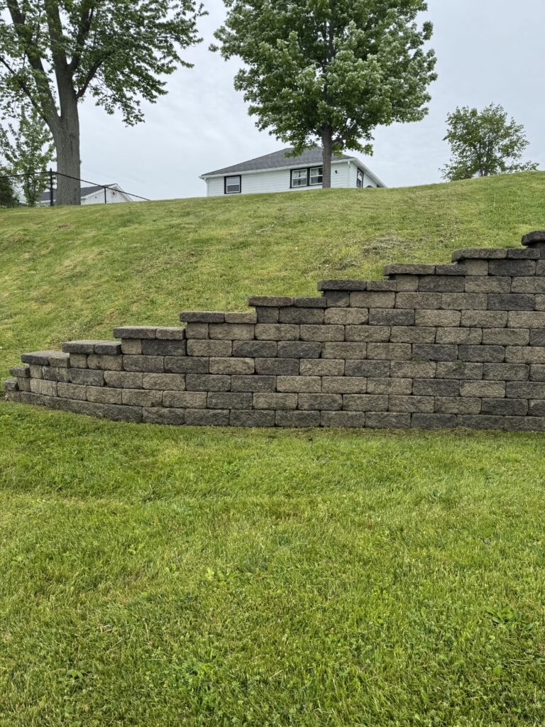 Tiered concrete block retaining wall along a grassy slope with a white house on a hill in the background and trees nearby.