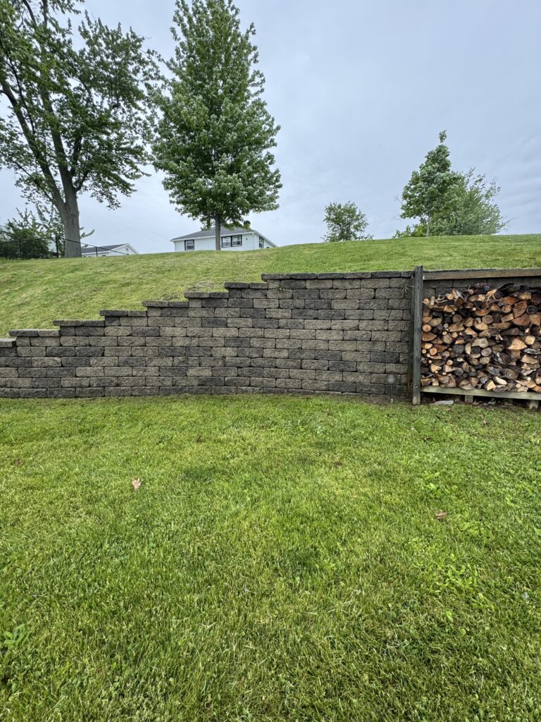 Grassy yard with a stacked stone retaining wall and woodpile on the right, a small white house behind trees in the distance.