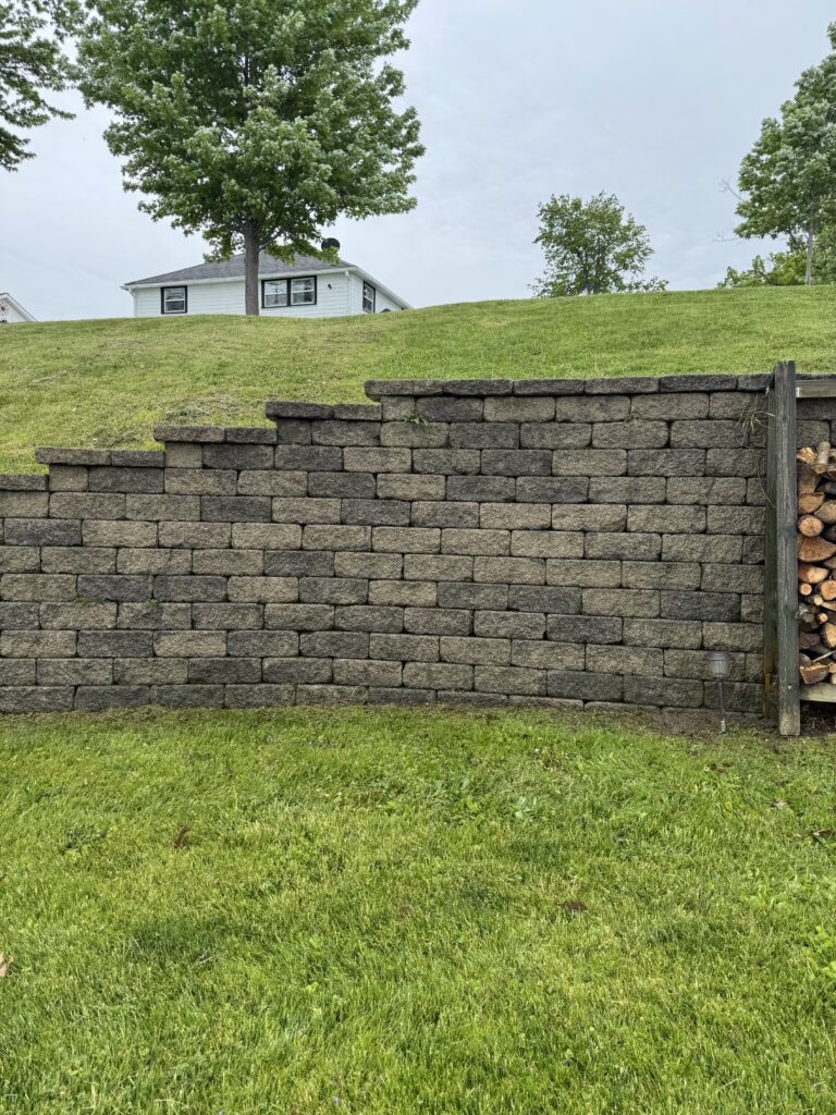Stone retaining wall in the foreground; green lawn and trees with a small white house on a hill in the distance.