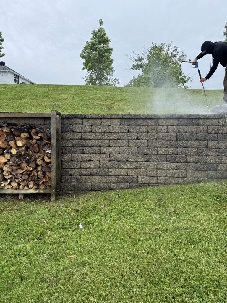 Person using a pressure washer to clean a stone retaining wall outdoors, with a wood pile and grassy slope nearby.