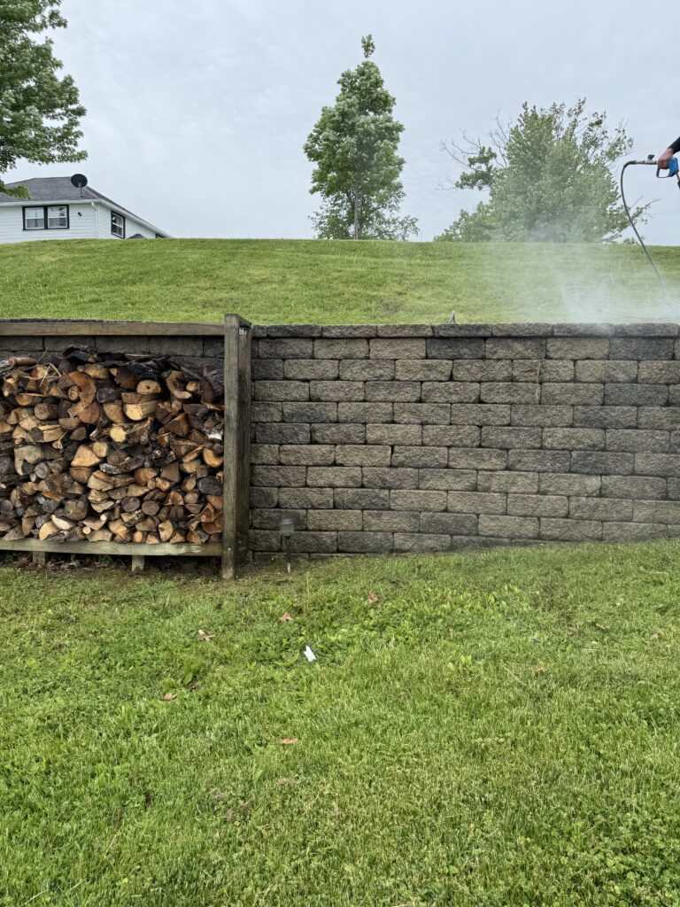 Pressure washer user spraying water mist over a stone retaining wall with a wood pile on the left and a grassy hill in the background.