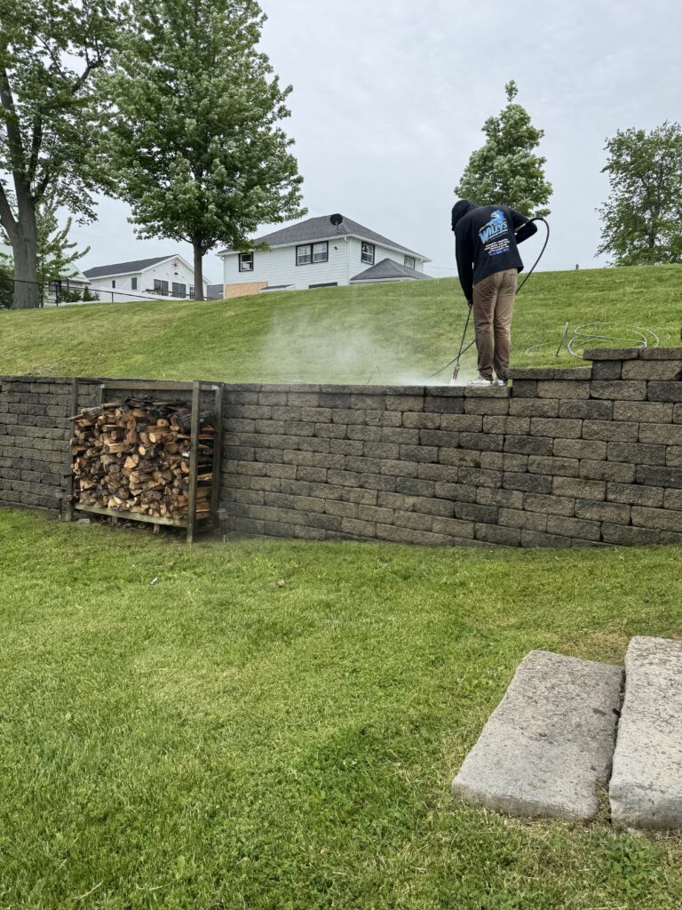 Person in a dark hoodie pressure-washing a stone retaining wall; mist rises from the nozzle with a grassy yard and houses in the background.