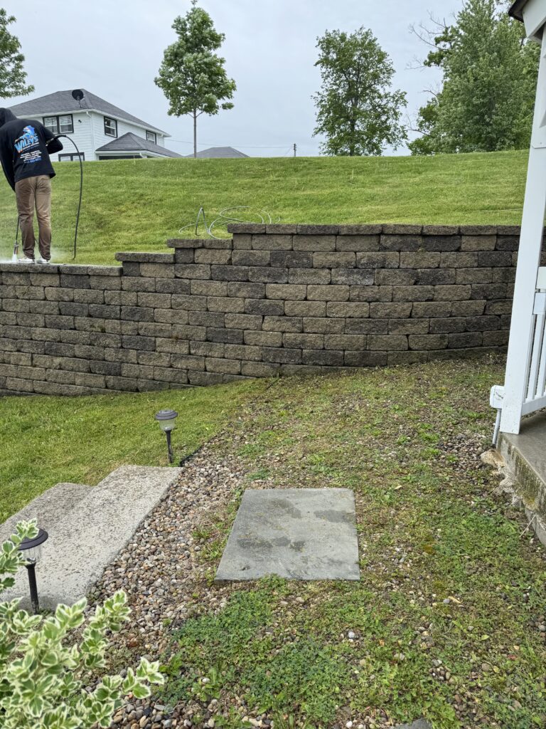 Stone retaining wall with stepped edge, gravel path, and small landscape lights in a backyard; a person in a dark hoodie stands on the wall.
