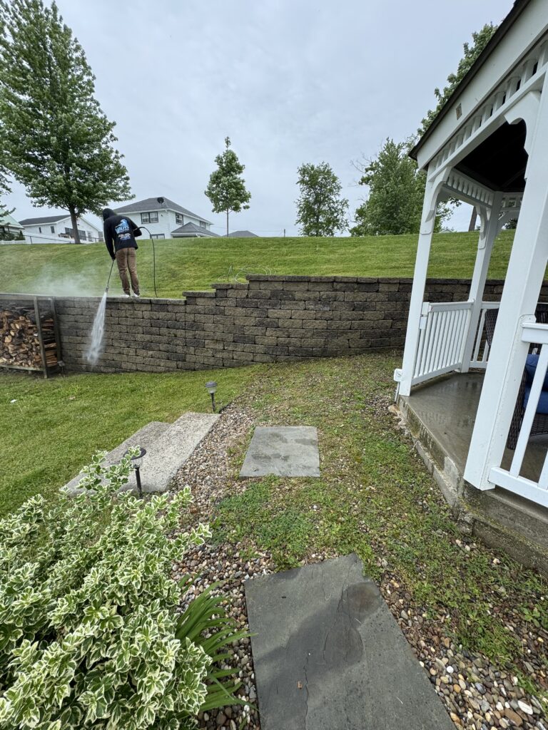Person in a dark hoodie using a pressure washer to clean a stone retaining wall in a backyard, water spray visible from the nozzle.