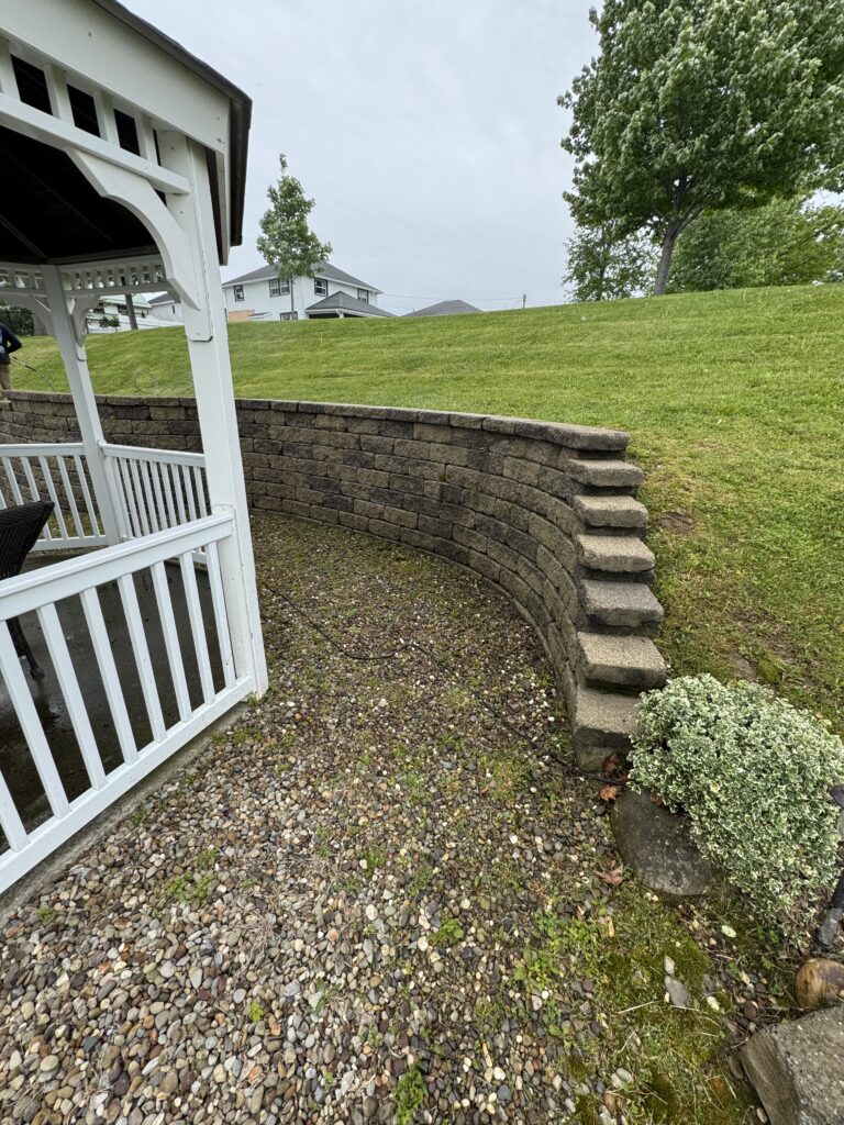 White porch railing with peg-style posts sits above a gravel/pebble patio, curved stacked-stone wall and a green grassy hill beyond with trees in the distance.