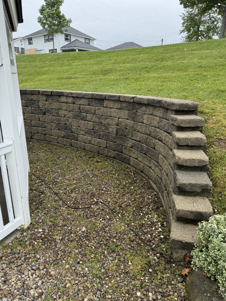 Curved stacked-stone retaining wall with built-in steps on the right, separating gravel foreground from a grassy hill.