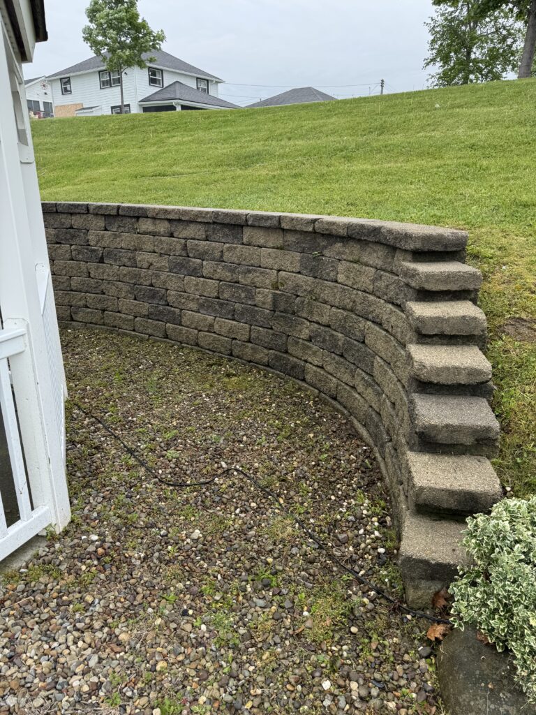 Curved gray block retaining wall with integrated steps along a gravel patio, separating a lower area from a grassy, sloped yard behind a house.