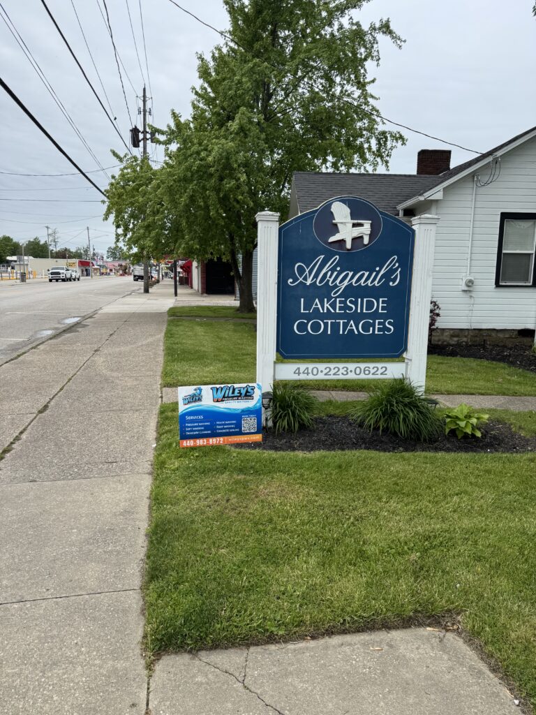 Blue sign for Abigail's Lakeside Cottages along a sidewalk with a large tree nearby, in a small-town street scene.