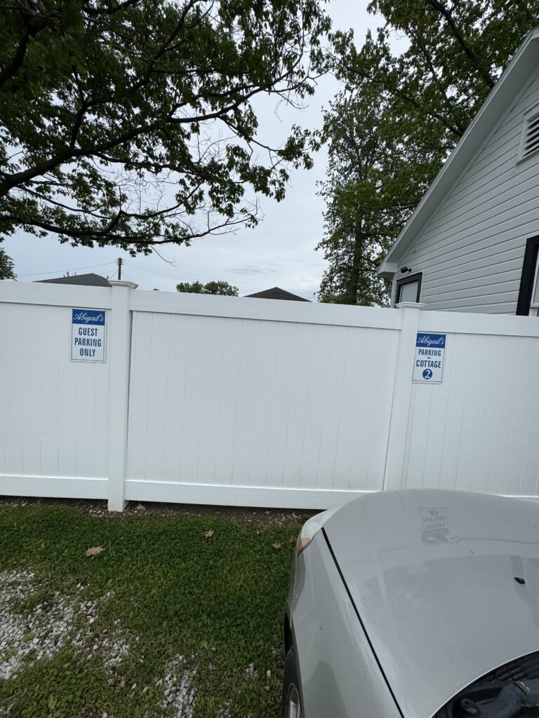 White privacy fence with two blue signs reading 'Abigail's Guest Parking Only' and 'Parking for Cottage 2', a gray car in foreground and tree branches overhead.