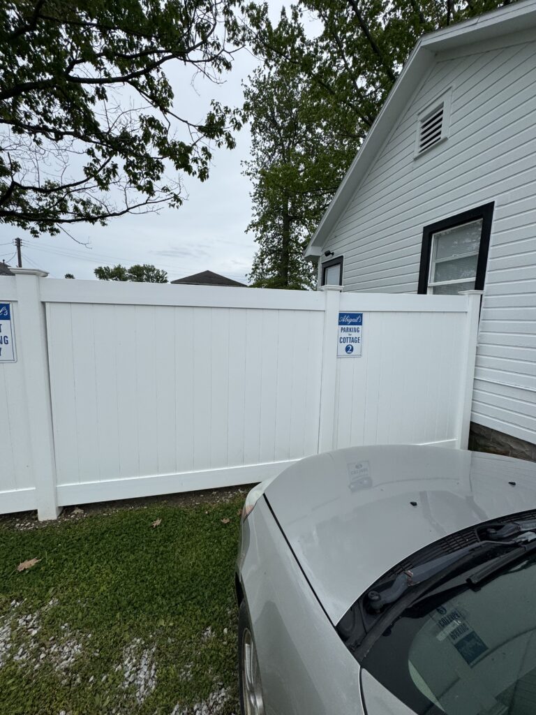 White vinyl privacy fence with a parked silver car in front, adjoining a white house with a vent and window, under leafy trees on an overcast day.