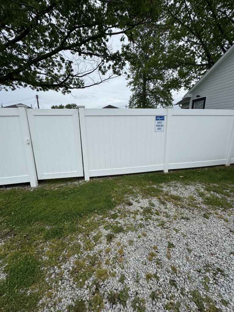 White privacy fence with a small gate, gravel driveway in front, and a large tree overhead; a sign on the fence reads 'Guest Parking Only'.