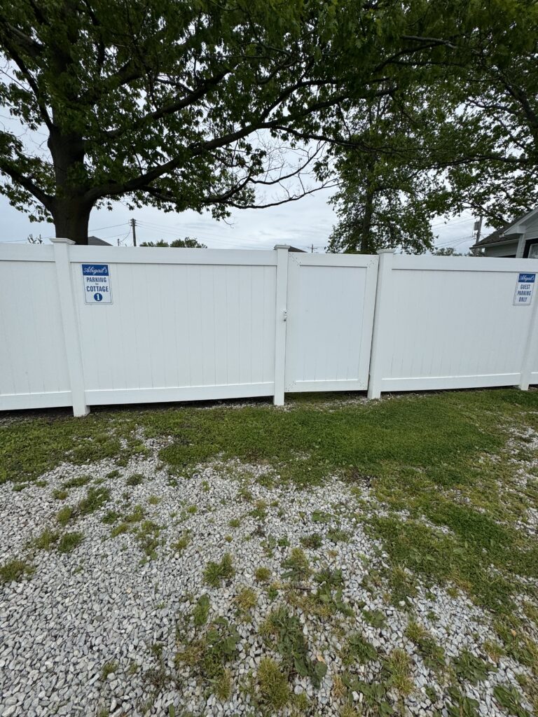 White vinyl fence with two blue parking signs ('Abigail's Parking Cottage 1' and 'Guest Parking Only'), gravel foreground, large tree behind.