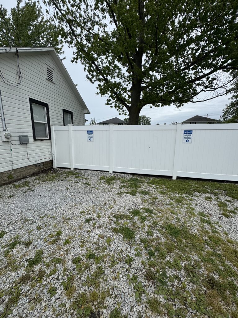 Side view of a white house with two windows, a white fence, and a large tree behind the fence over a gravel yard with weeds.