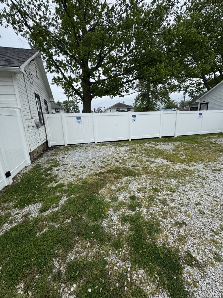 White vinyl fence enclosing a yard with a large tree and gravel ground, houses visible beyond the fence.