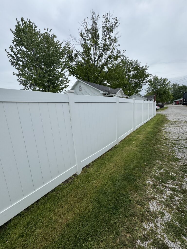 White vinyl privacy fence runs along a grassy edge beside a gravel driveway with trees and houses in the background under a cloudy sky.] ,