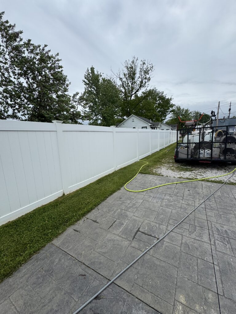 Backyard scene with a tall white fence, green grass, and a trailer carrying equipment on the right under a cloudy sky