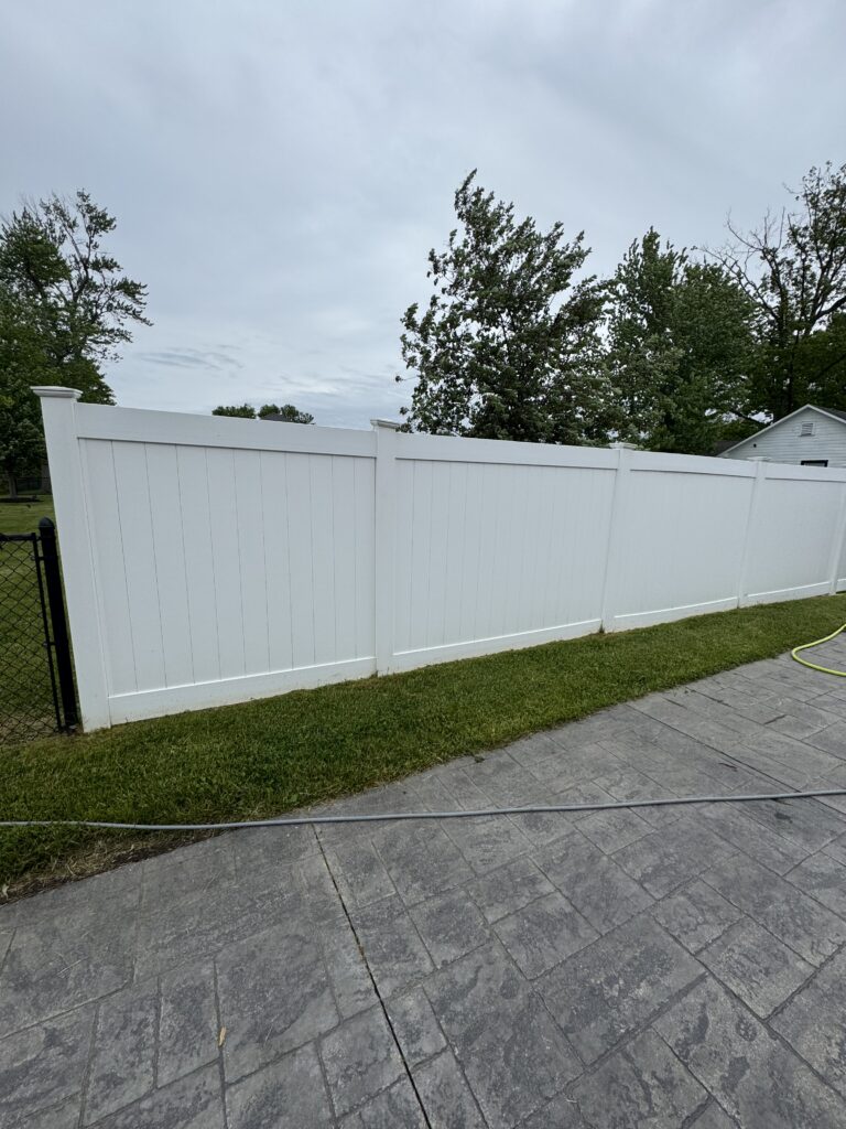 White vinyl privacy fence runs along a grassy yard with a gray paved patio; a yellow garden hose lies on the ground, trees and a cloudy sky in the background.