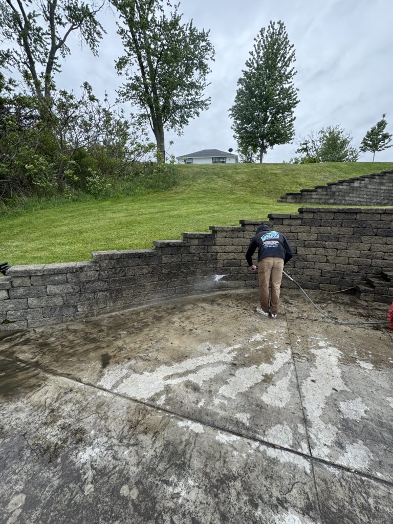 Person in a hoodie power-washing the inside of a multi-tiered stone wall, with a muddy concrete area at his feet and a grassy slope behind him.