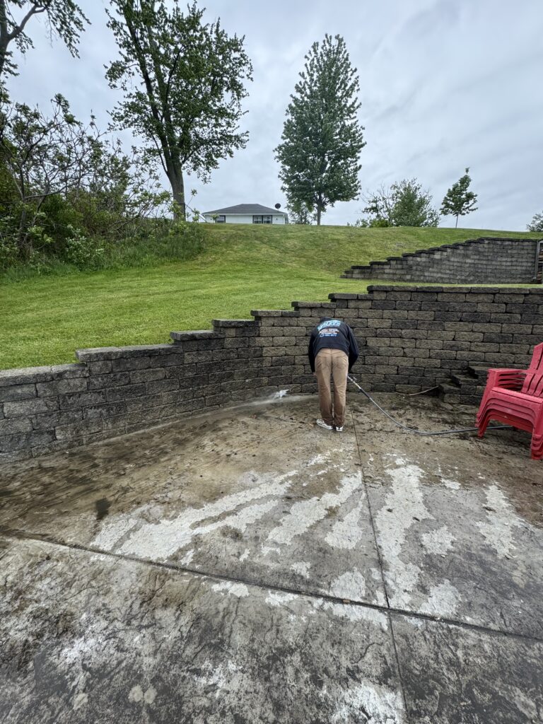 Person bending over and power-washing a concrete patio near stone retaining walls, with a grassy hill and trees in the background and red chairs on the side.
