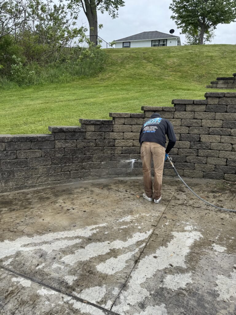 Worker in a dark jacket pressure-washing a stacked stone retaining wall by a grassy slope
