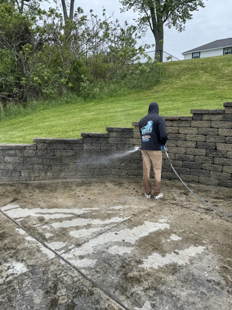 Person in a dark hoodie using a pressure washer to clean a stacked stone retaining wall by a grassy yard.