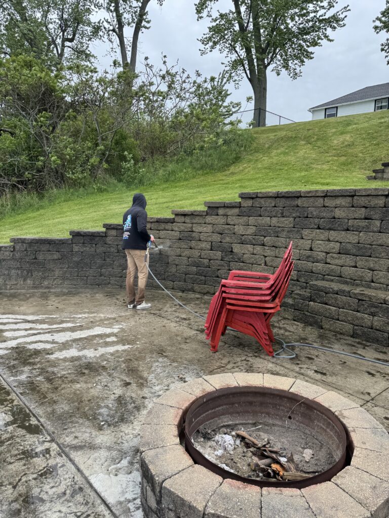 Person watering a garden area beside a circular brick fire pit and stacked red chairs on concrete patio.