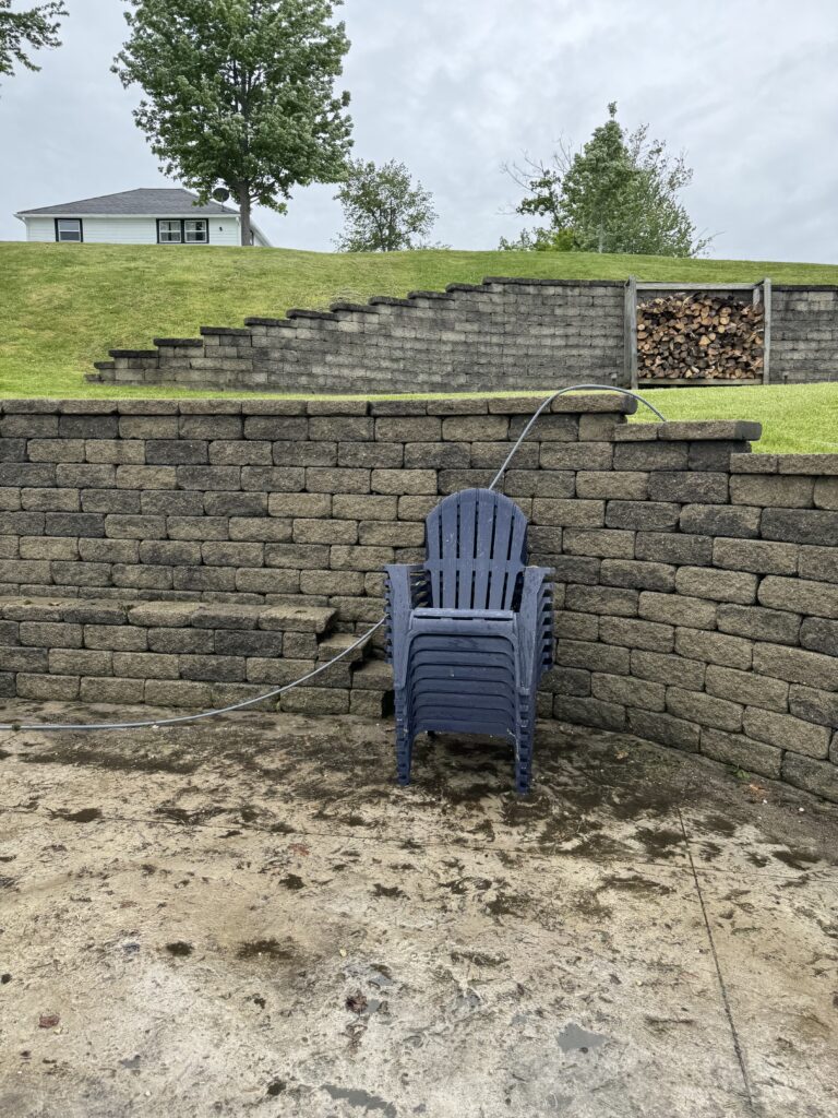 Stacked stone retaining wall with blue plastic Adirondack chairs stacked in front, hose running over wall, muddy ground in foreground.