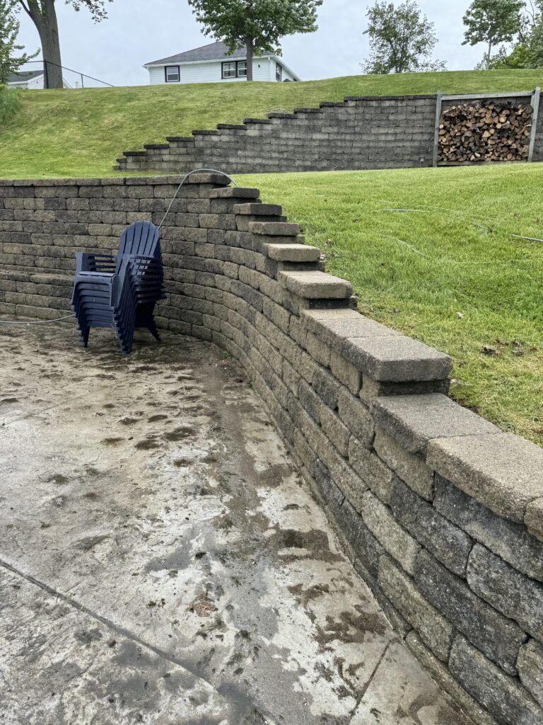 Stack of blue plastic chairs leaning against a curved metal rod beside a concrete block retaining wall in a wet patio area with a grassy hill behind it.