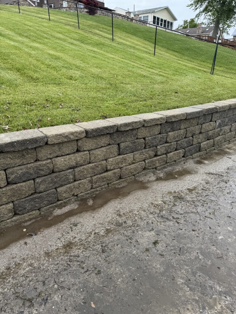 Stone tiered retaining wall beside a wet driveway, with a green sloped lawn and chain‑link fence in the background.