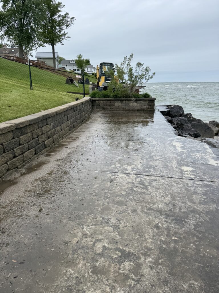 Waterfront park with a stone retaining wall, wet concrete path, and a yellow excavator by the shore among trees and houses nearby