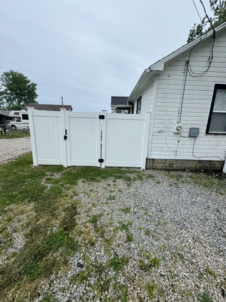 White wooden privacy gate beside a white house with a gravel yard and sparse grass growth behind it.