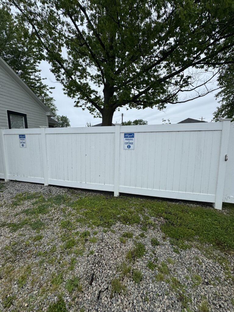 White wooden privacy fence with blue parking signs, cottage numbers 1 and 3, in front of a yard with a large tree behind it