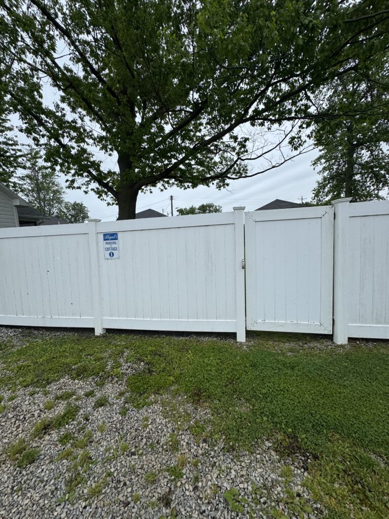 White wooden privacy fence with a gate, a large tree behind it, and a small sign on the fence reading 'Parking Cottage'; gravel and weeds on the ground in the foreground, cloudy sky above.