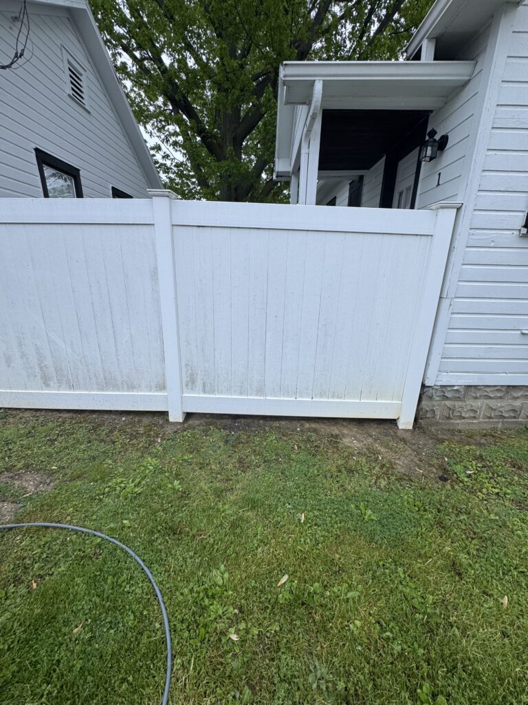 White wooden backyard gate in front of a white house, with a large green tree behind and a garden hose on the grass in the foreground.
