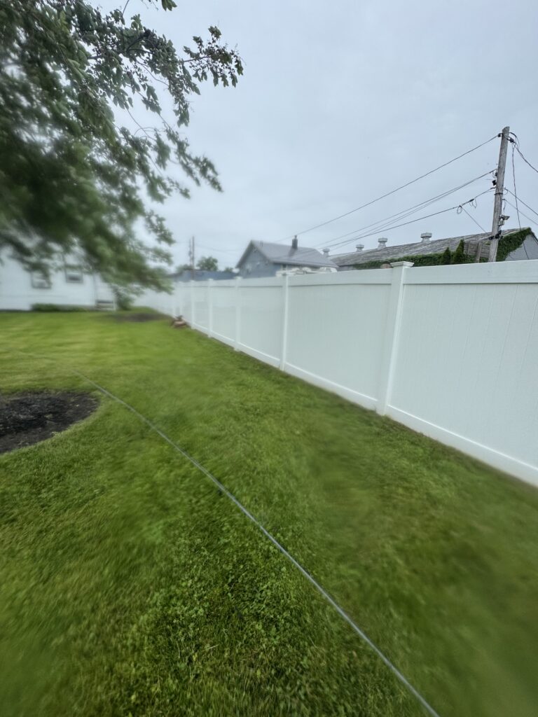 Backyard scene with a tall white fence, green lawn, and overhanging tree branches on the left under an cloudy sky, with a utility pole and power lines in the background.