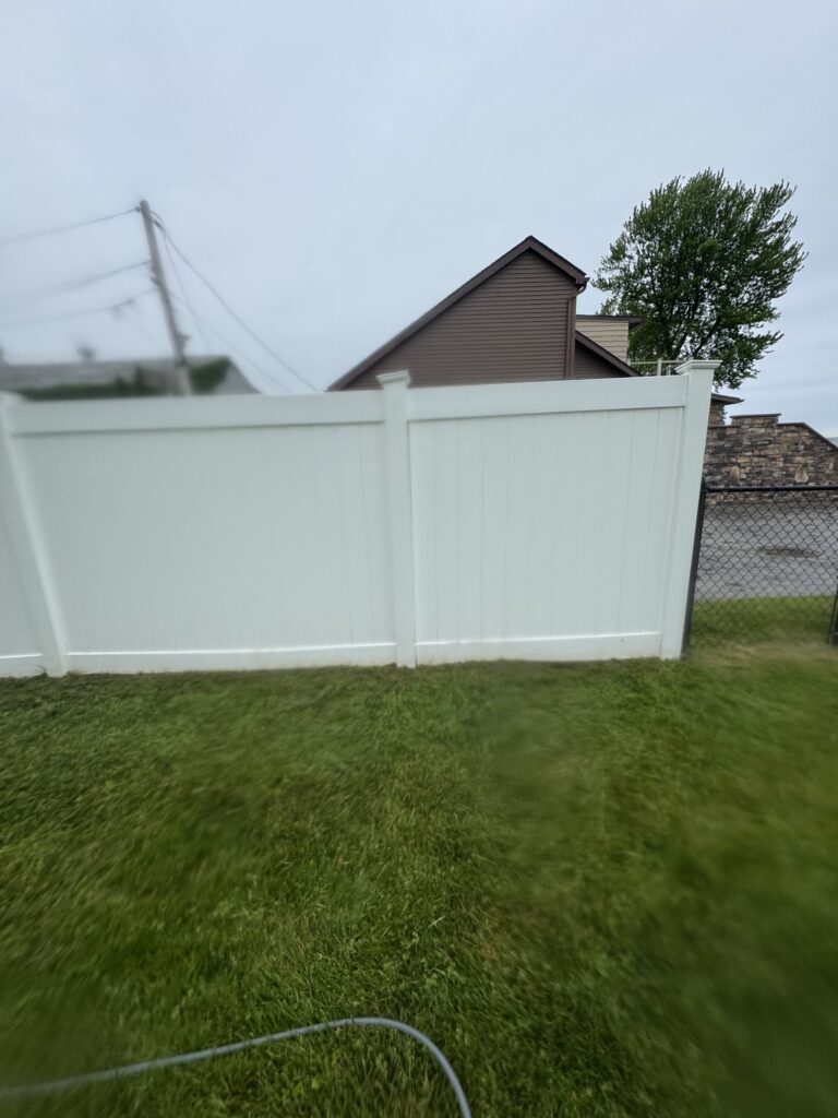 White privacy fence in the foreground with a brown peaked-roof house behind, plus a tree and chain-link fence in a grassy yard.
