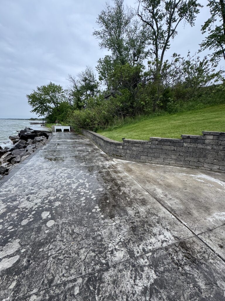Overcast waterfront concrete walkway along a rocky shore with a grassy slope and trees behind a stone wall, by the water.