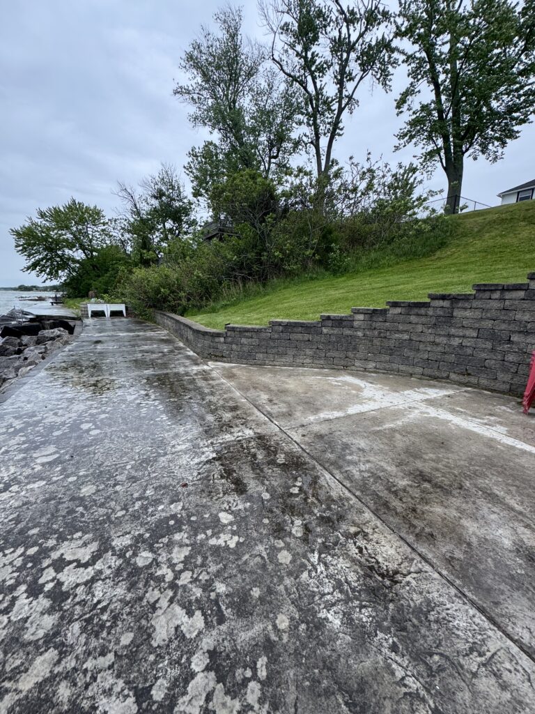 Concrete waterfront walkway with a stone retaining wall, green grassy slope, and tall trees under a cloudy sky by the water's edge