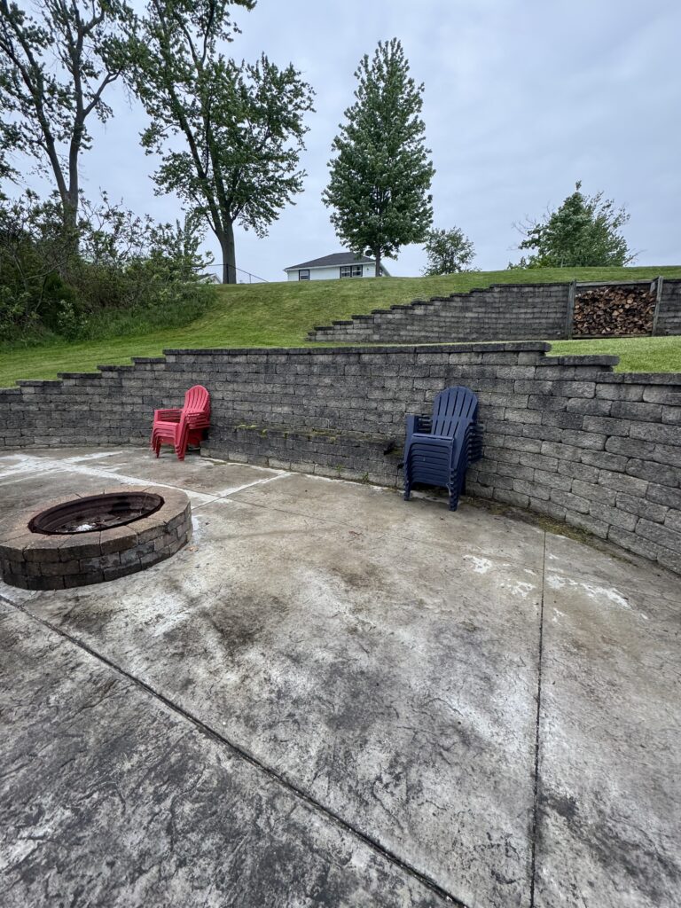 Outdoor patio with circular fire pit, stacked stone seating, and red and blue plastic chairs on a concrete surface; a grassy slope and trees in the background.