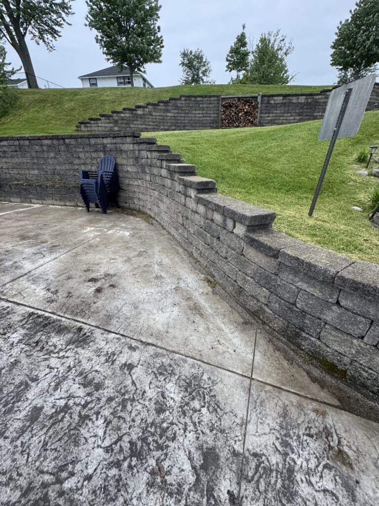 Backyard with gray stacked-block retaining walls in a tiered terrace and stacked blue plastic chairs at the base of the wall.
