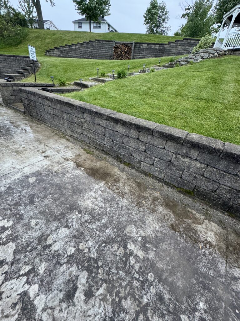 Stone retaining walls and a sloped grassy yard with a concrete path in the foreground and a small sign on the left edge of the scene.