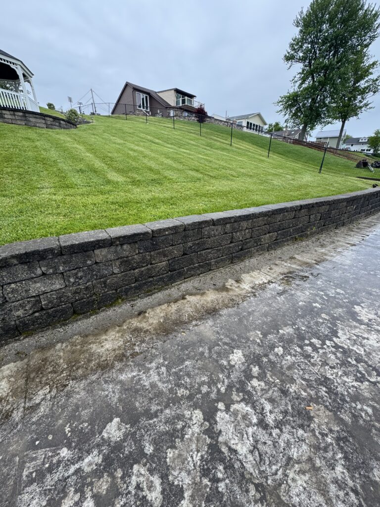 Sloped, well-manicured green lawn with a stone retaining wall and fence, houses in the distance under an overcast sky.