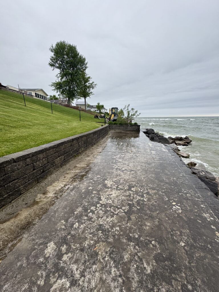Seaside walkway along a stone seawall with a sloped grassy hill, trees, and houses in the distance under a cloudy sky.
