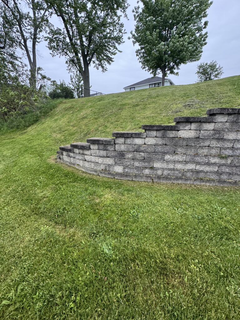 Sloped grassy yard with a stacked stone retaining wall in the foreground and a white house atop the hill among several trees