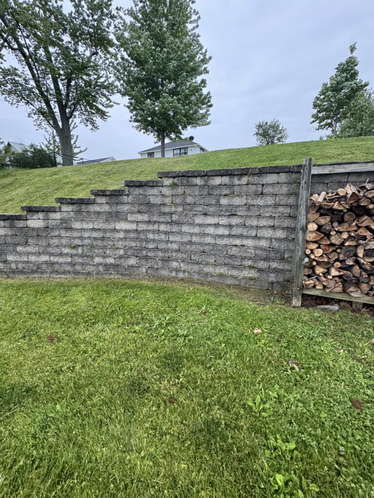 Backyard scene with a multi-layer stone retaining wall, a green slope, and a stack of firewood on the right; trees and a house are in the distance.